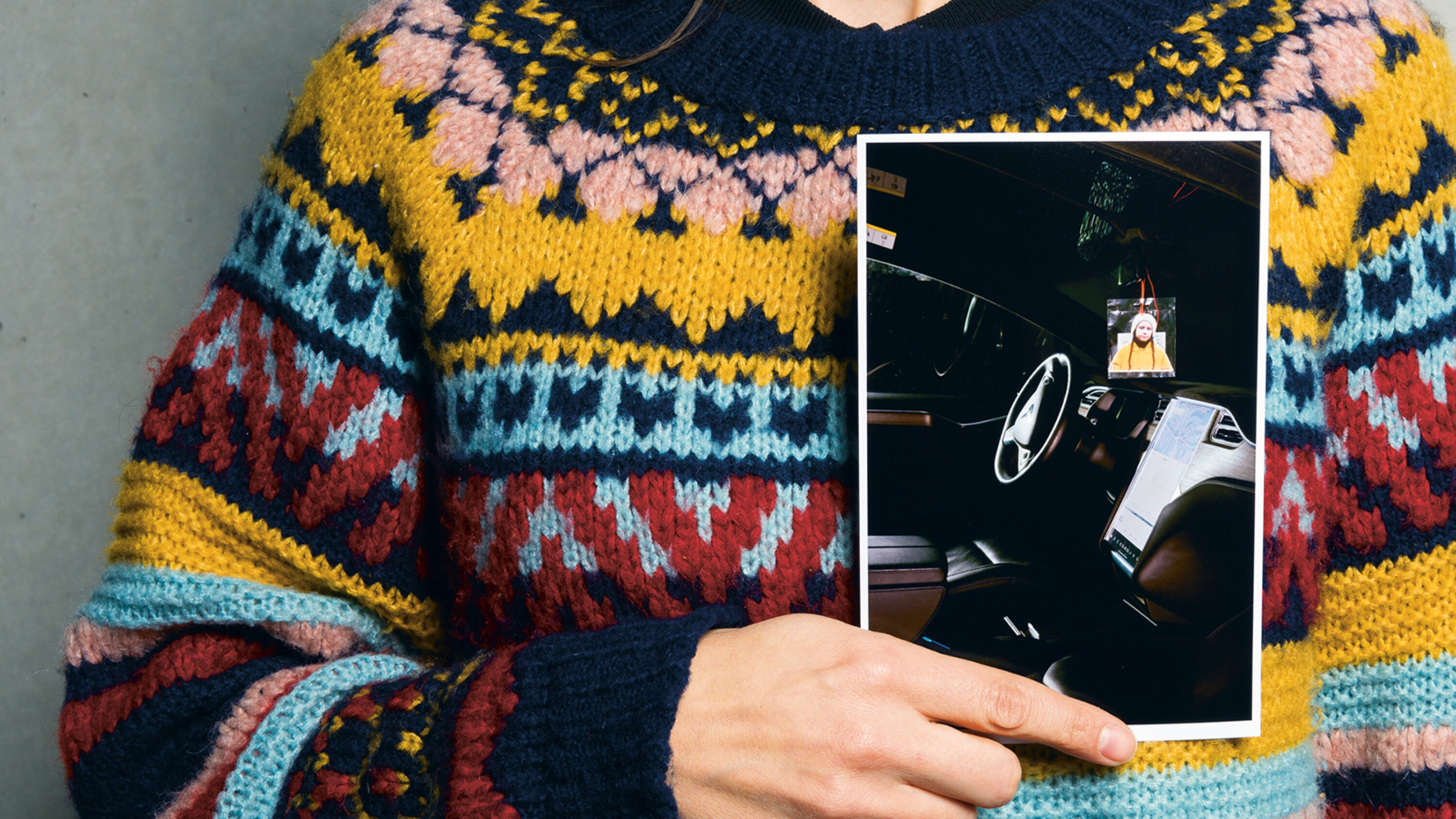 Person in gestricktem Pullover zeigt ein Foto mit Autoinnenraum und aufgehängtem Porträt.