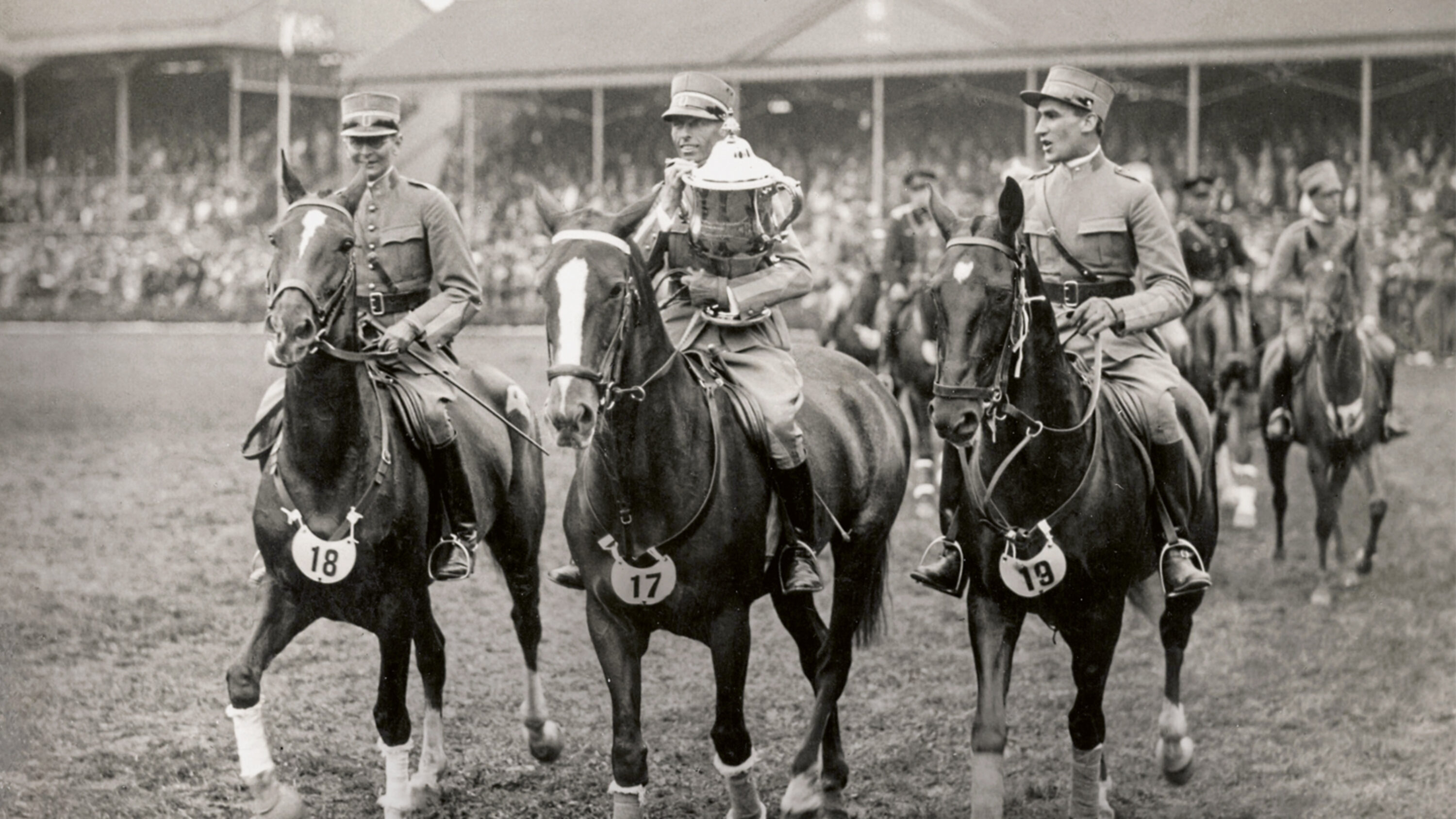 Drei Reiter in Militäruniform bei einem historischen Pferdesportwettbewerb im Stadion.