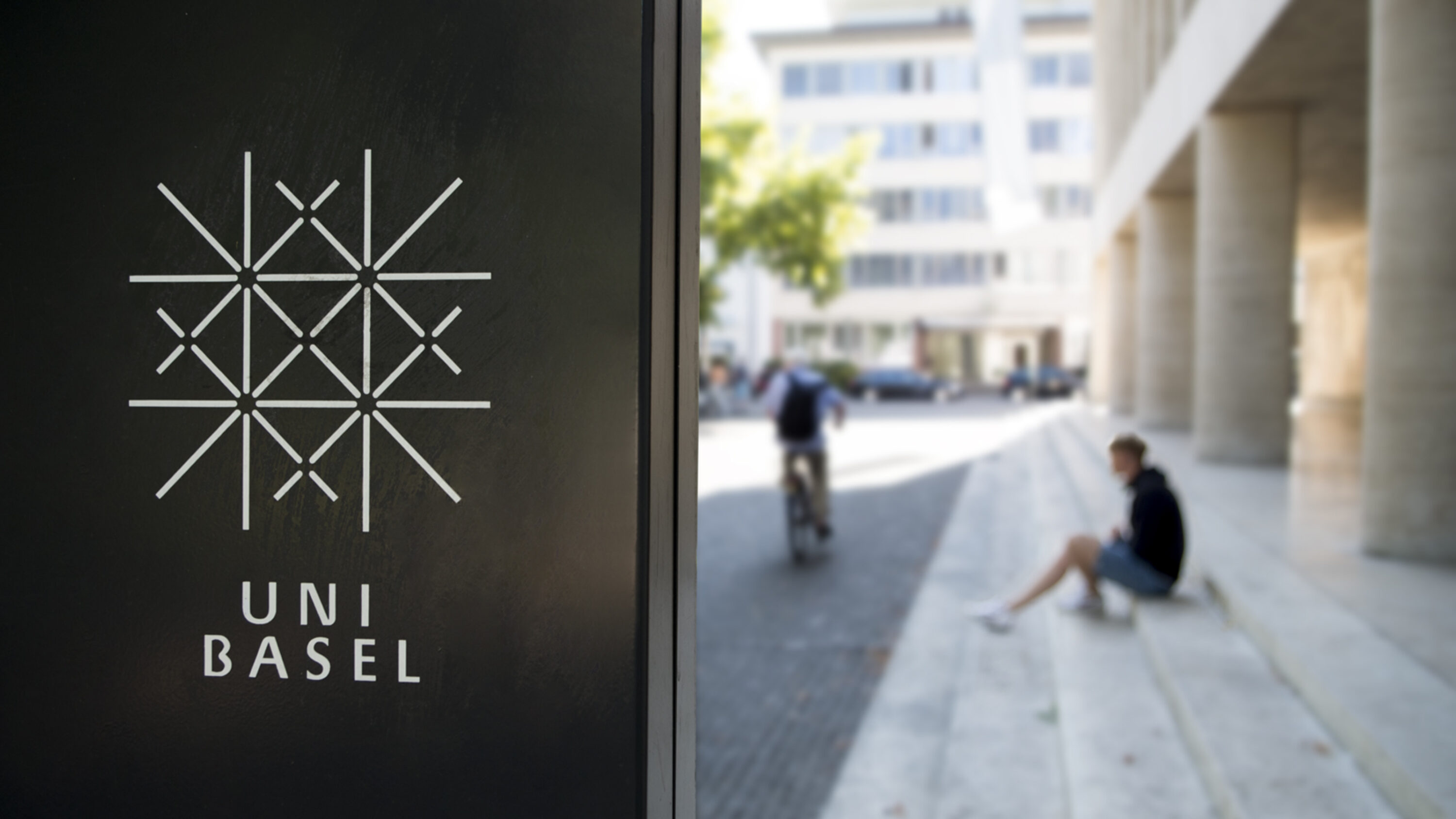 Logo der Universität Basel auf einer dunklen Tafel im Vordergrund, im Hintergrund unscharf ein Campusbereich mit einem sitzenden Mann und einer Person auf einem Fahrrad.